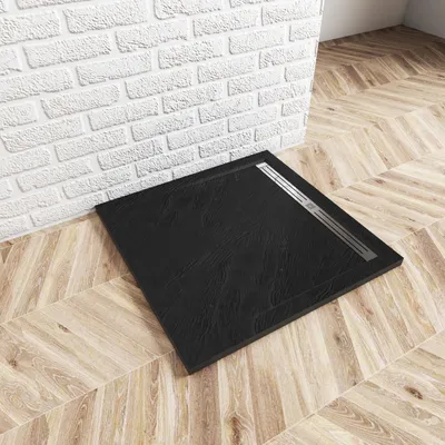 A black square shower tray with a silver drain cover is placed on a wooden floor next to a white brick wall.