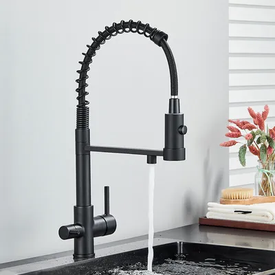 A black kitchen faucet with a pull-down sprayer is shown in use, with water flowing into a sink. The background is a plain wall and a window with blinds. A small vase with dried flowers and a brush are visible on a tray to the right.