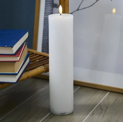 A white LED candle stands on a wooden floor next to a stack of books and a framed picture.