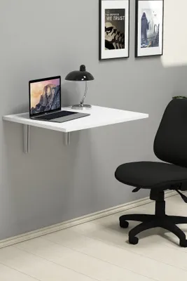 A white folding desk mounted on a gray wall, with a laptop and a lamp on it. A black office chair is in the foreground.