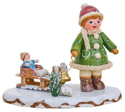 Ein fröhliches Winterkind in grüner Kleidung steht auf einer schneebedeckten Basis neben einem Schlitten mit einem sitzenden Kind und einem kleinen Weihnachtsbaum.