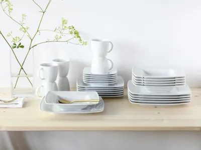 A collection of white porcelain dinnerware, including plates and mugs, neatly stacked on a wooden shelf against a white background.