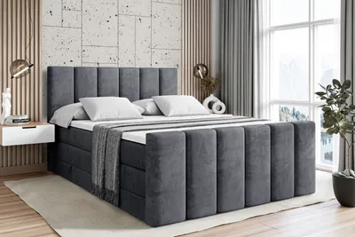 A dark gray upholstered box spring bed with a tufted headboard and footboard, set up in a bedroom with a bedside table and a large window in the background.