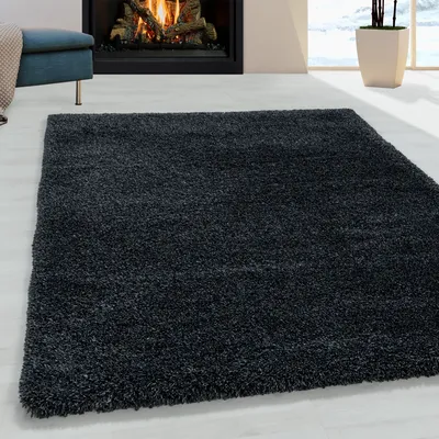 A dark shaggy rug is placed on a light-colored floor in front of a fireplace. A blue ottoman with a beige throw is visible to the left. A potted plant is in the background to the right.