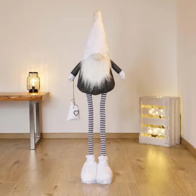 A decorative gnome with long legs stands in a room with a table and lamp on the left and a wooden crate with lights on the right.