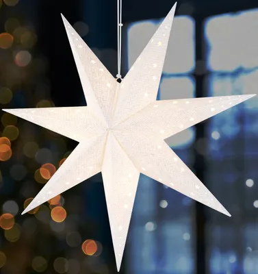A white paper star lantern with small white dots is hanging in front of a window with blurred lights in the background.