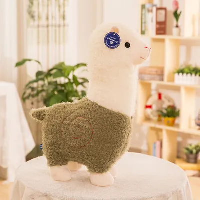 A plush alpaca toy is displayed on a table with a blurred background of shelves and plants.