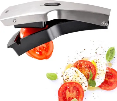 A tomato slicer is shown in action, slicing a tomato. Sliced tomatoes and mozzarella cheese are arranged in the background.