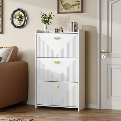 A white shoe cabinet with three flip-down doors and gold handles is shown in a room next to a brown sofa and a door.