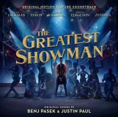 The image is a promotional poster for the soundtrack of the movie "The Greatest Showman". It features the movie title prominently, along with the names of the main actors and the original songwriters. The background depicts a circus-like scene with spotlig