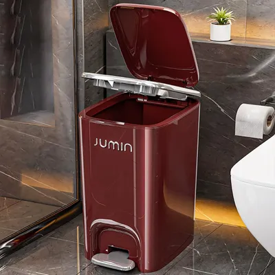 A red pedal trash can with the lid open, placed in a bathroom next to a toilet and a roll of toilet paper.