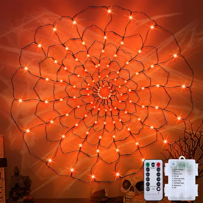 A large, illuminated spider web decoration with orange lights, set against a dark, possibly orange-tinted background, with some faux cobwebs. A remote control and battery pack are visible in the lower right corner, along with a skull decoration.
