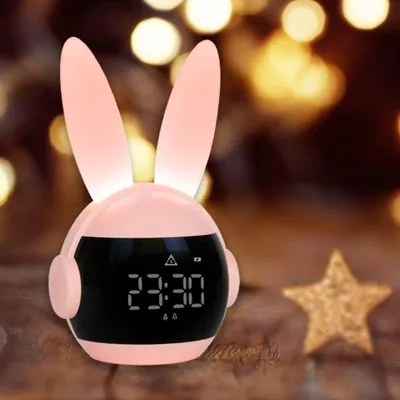 A pink bunny-shaped alarm clock with a digital display and glowing ears sits on a wooden surface, with blurred bokeh lights in the background.