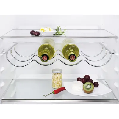 Inside a refrigerator, a metal wine rack holds two bottles of white wine. Below, a shelf displays a small jar of corn, a red chili pepper, grapes, kiwi, and cheese.
