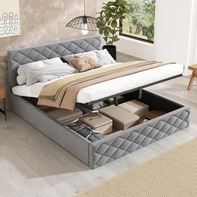 A grey upholstered bed with its storage compartment open, revealing stored items. The bed is made of velvet fabric and has a diamond-stitched headboard. It is set in a room with light wooden flooring and a large window in the background.