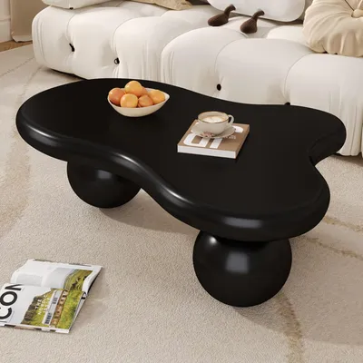 A black, cloud-shaped coffee table with a bowl of fruit and a cup of coffee on it, placed on a carpet in front of a white sofa.