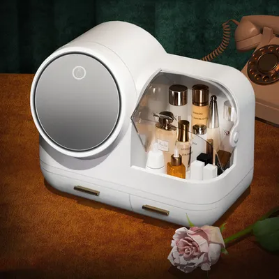 A white makeup organizer with a mirror and drawers, filled with various cosmetic products, sits on a brown surface next to a pink rose. A vintage telephone is visible in the background.