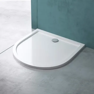A white, quarter-circle shower tray with a chrome drain is shown in a bathroom setting with concrete-like flooring and teal walls.