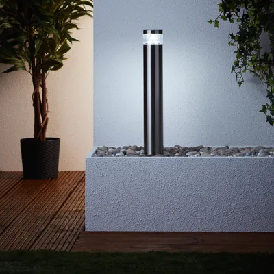 A modern outdoor lighting fixture is shown in a garden setting, next to a plant and a planter filled with stones.