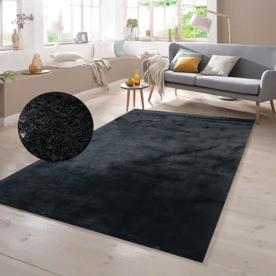 A dark gray shaggy rug is placed on a light wooden floor in a living room setting. The rug is in the foreground, with a gray sofa and a small side table visible in the background. Natural light streams in from large windows.