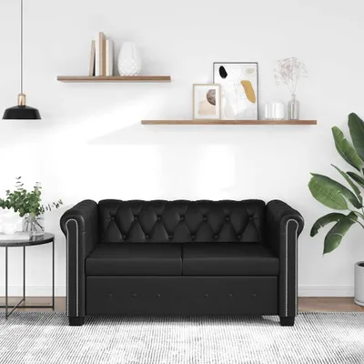 A black Chesterfield sofa is shown in a well-lit room with shelves on the wall.