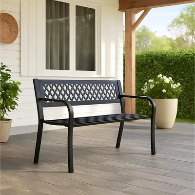 A black metal garden bench is placed on a wooden deck in front of a white house with windows. A large potted plant with white flowers is to the right of the bench, and a green hedge is in the background.