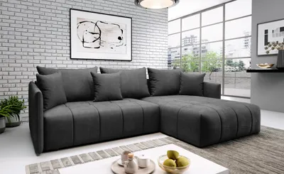 A modern, dark gray sectional sofa with multiple throw pillows is displayed in a living room setting. The sofa is positioned in front of a white brick wall with a piece of abstract art hanging above it, and large windows in the background that offer a view