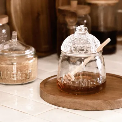 A honey jar with a dipper on a wooden tray, next to a jar of sugar.