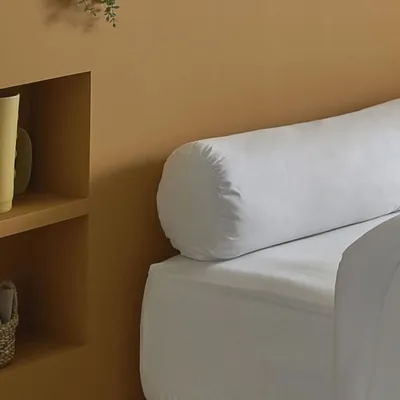 A white bolster pillow on a white bed against a tan wall.