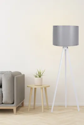 A floor lamp with a gray shade and white tripod base stands next to a gray sofa and a small wooden side table with a potted plant.