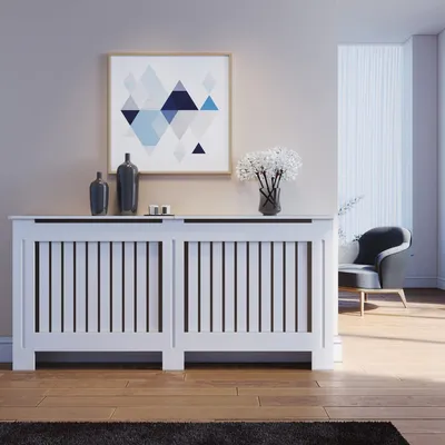 A white radiator cover with vertical slats is placed in a room with a framed abstract geometric artwork above it. Decorative vases and a bouquet of flowers are on top of the cover. A chair and a window with blinds are visible in the background.