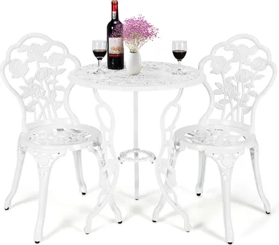 A white metal bistro set with a round table and two chairs, decorated with rose motifs. A bottle of red wine, two glasses of red wine, and a small vase with pink flowers are placed on the table.