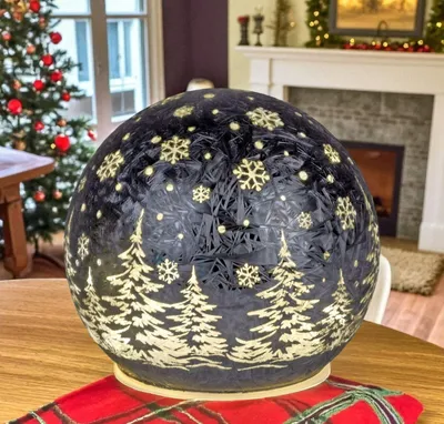 A decorative LED sphere with a Christmas theme is placed on a table in a room with a Christmas tree and fireplace in the background.