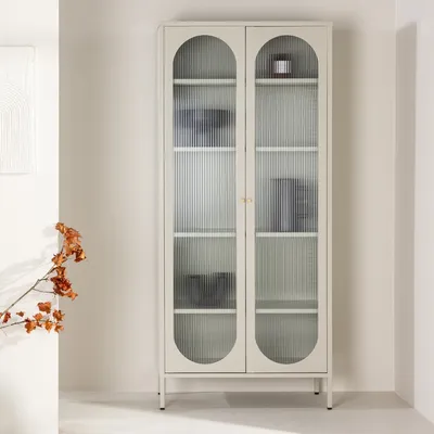 A beige display cabinet with fluted glass doors and arched tops, standing in a minimalist room with a branch of dried flowers to the left.
