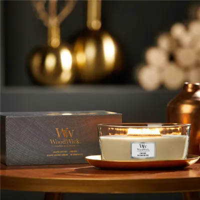 A WoodWick candle and its packaging are displayed on a wooden table with decorative golden objects in the background.