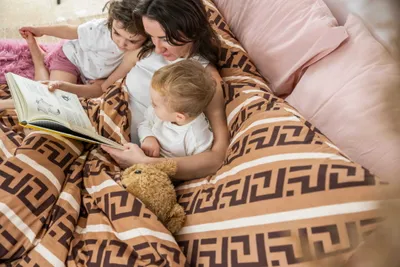 A mother reads a book to her two young children in bed, under a patterned blanket.