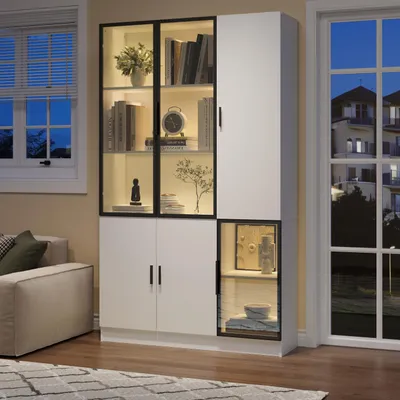 A modern white vitrine with glass doors and LED lighting, showcasing decorative items and books, placed in a living room setting next to a window.