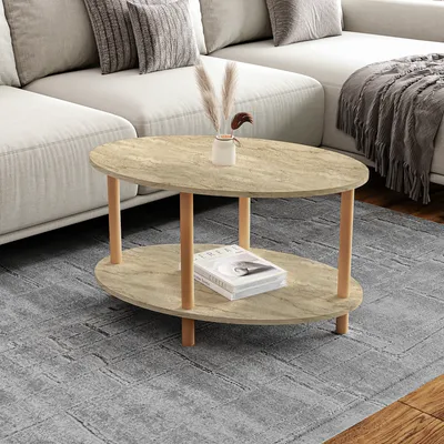 A modern living room scene featuring a two-tiered oval coffee table with a travertine-like finish and wooden legs, placed on a textured gray rug in front of a sectional sofa.