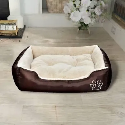 A brown and cream dog bed is placed on a light-colored wooden floor in a room with a fireplace and flowers in the background.