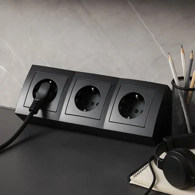 A black power strip with three outlets is shown on a desk, with a plug inserted into one outlet. Headphones and a notebook are also visible.