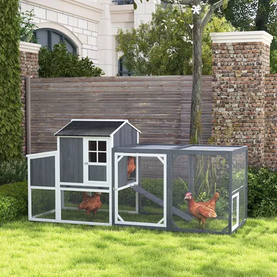 A wooden chicken coop with an attached run is set up on a grassy lawn in front of a wooden fence and a brick wall. Two chickens are visible inside the coop and run.