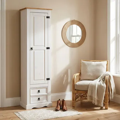 A tall white cabinet with a wooden top and two drawers is shown next to a wicker chair with a cushion and a blanket. A round mirror hangs on the wall behind the chair. Two brown boots are placed on a rug in front of the cabinet.