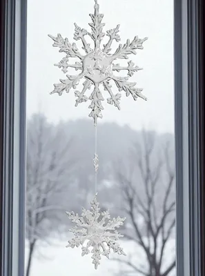 Two snowflake ornaments hang in a window with a snowy landscape visible outside.