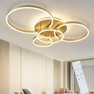 A modern gold ceiling lamp with multiple rings and integrated LED lights, shown in a well-lit room with a remote control in the foreground.