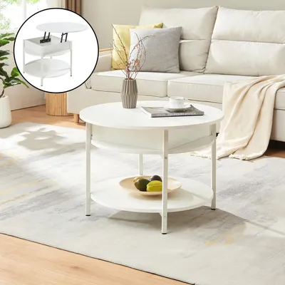 A white round coffee table with two shelves is placed on a light gray and white patterned rug in a living room setting. A sofa with cushions and a throw blanket is visible in the background.