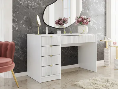 A white vanity desk with fluted drawers and gold handles, a pink chair, and decorative items on top, set against a textured gray wall.