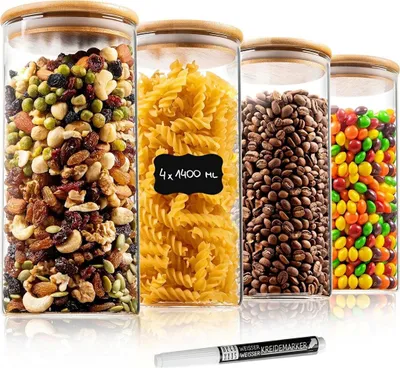 Vier klare Glas-Vorratsgläser mit hellen Bambusdeckeln, gefüllt mit Studentenfutter, Pasta, Kaffeebohnen und bunten Bonbons, mit einem weißen Kreidestift davor