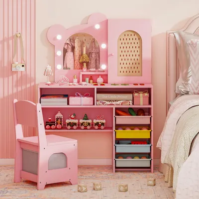 A child's pink vanity desk and chair set with various toys and accessories, placed in a bedroom setting next to a bed.