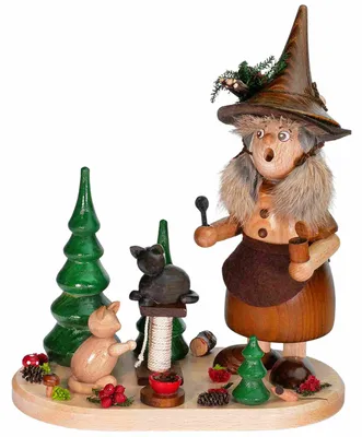 Holzfigur einer Wichtelfrau mit spitzem Hut, grauem Haar und braunem Kleid, umgeben von grünen Tannen, einer schwarzen Katze und roten Beeren auf rundem Holzsockel. Erzgebirgskunst aus Holz.