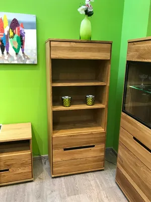 A wooden shelf unit with drawers and open shelves, decorated with a vase of flowers and two small decorative items. A painting hangs on the green wall behind it. A small wooden side table is to the left.